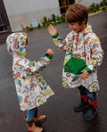 Duas crianças sorridentes, de costas e de frente, a usar ponchos Hello Hossy San Francisco coloridos em dia de chuva, brincando ao ar livre.