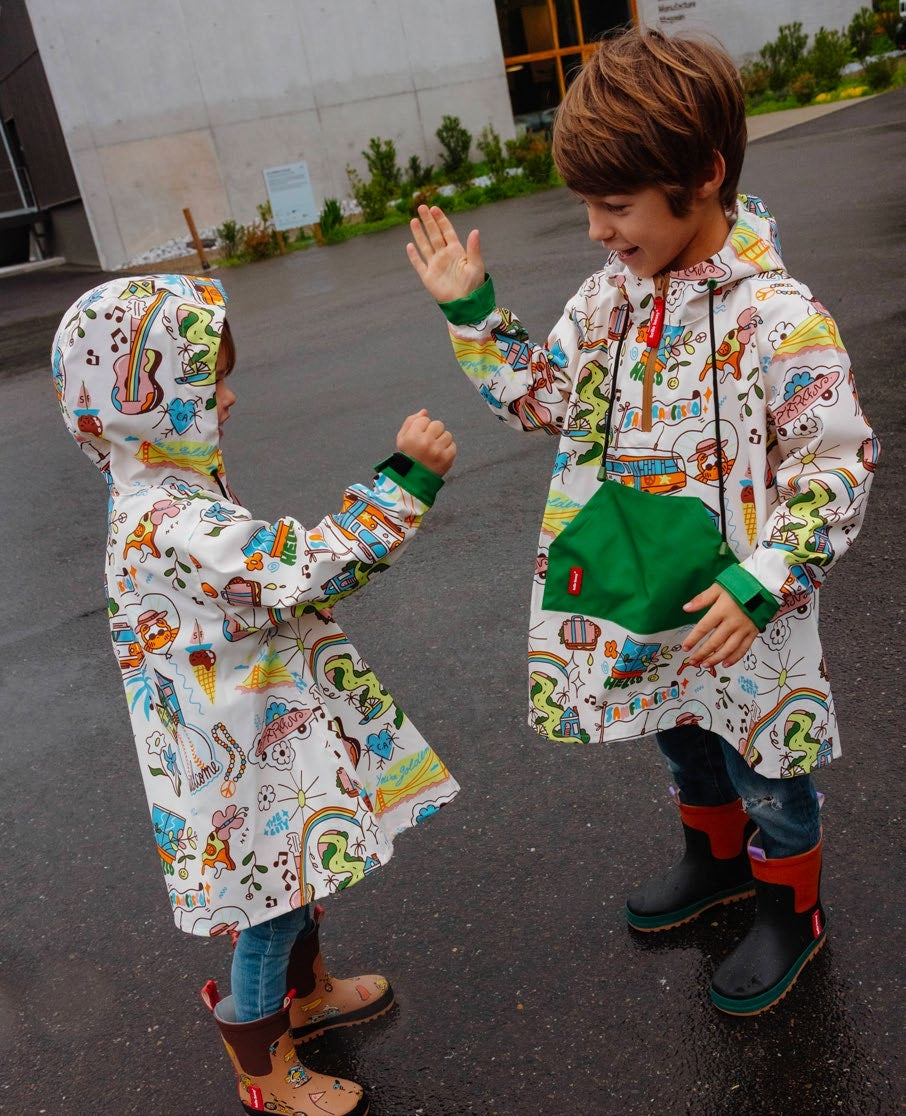 Duas crianças sorridentes, de costas e de frente, a usar ponchos Hello Hossy San Francisco coloridos em dia de chuva, brincando ao ar livre.
