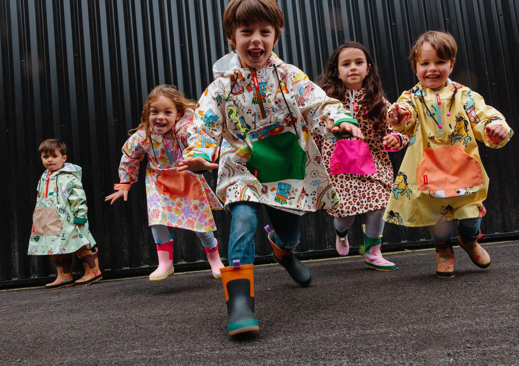Crianças alegres a correr, vestindo vários modelos de ponchos impermeáveis Hello Hossy com padrões coloridos, contra um fundo escuro.