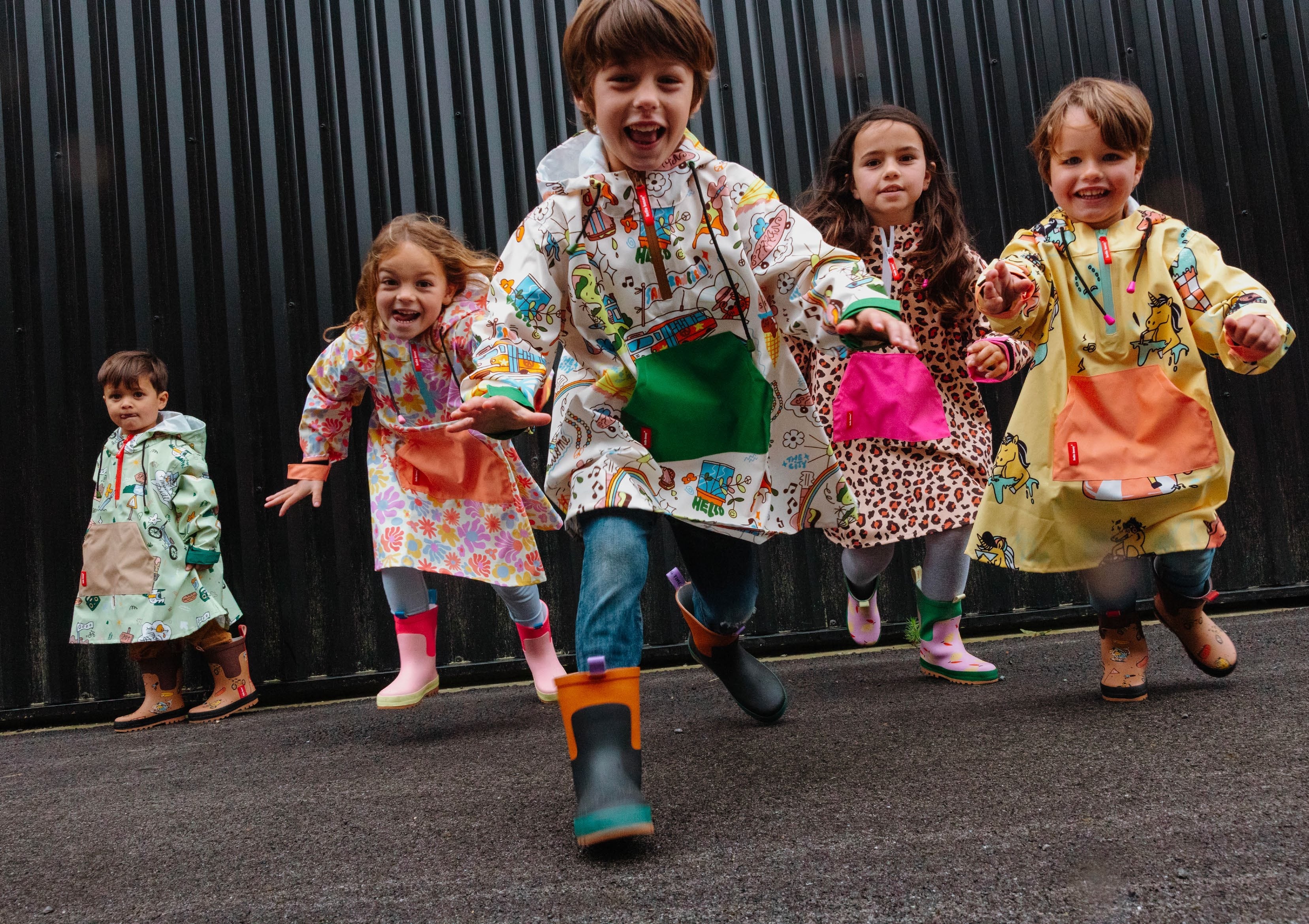 Crianças alegres a correr, vestindo vários modelos de ponchos impermeáveis Hello Hossy com padrões coloridos, contra um fundo escuro.