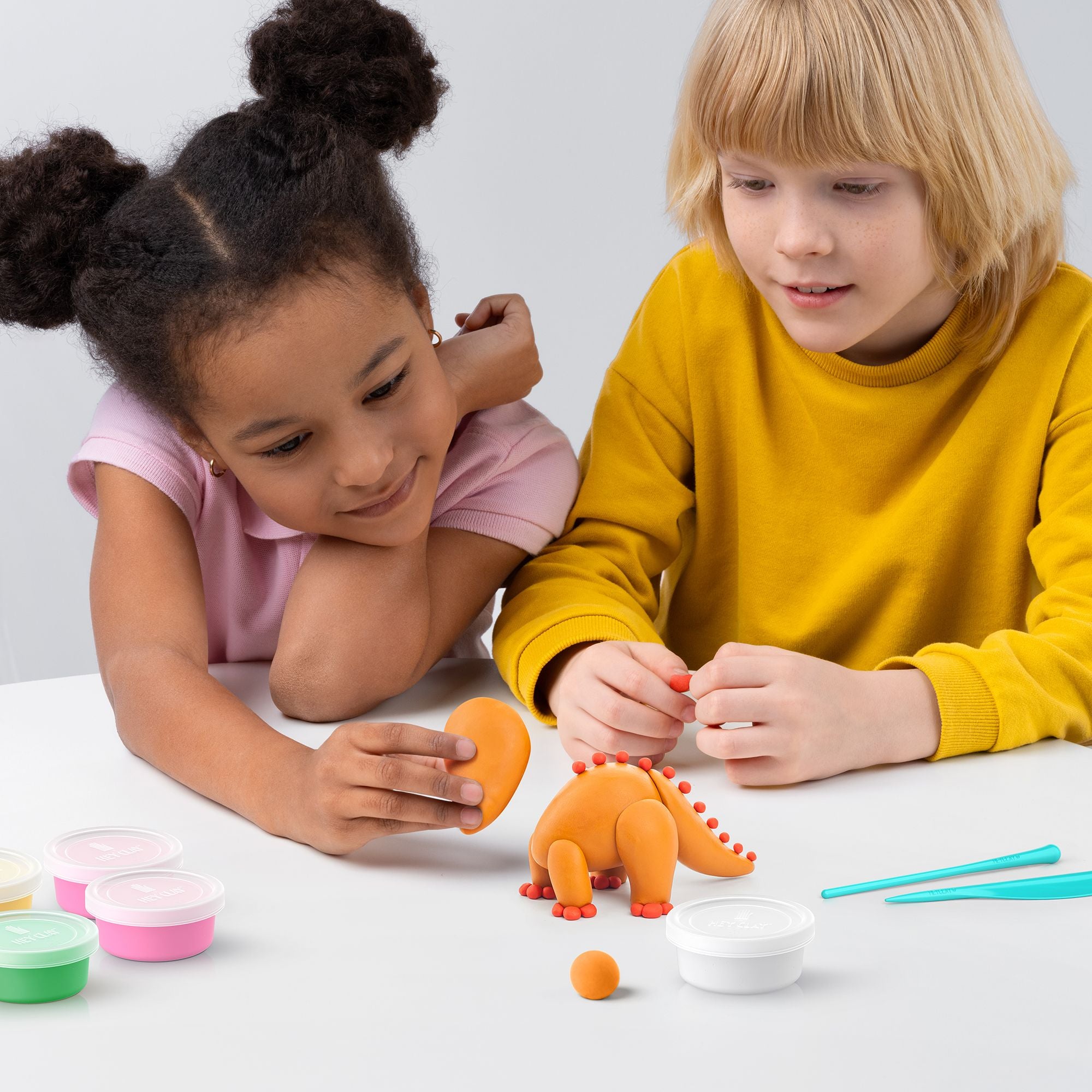 Duas crianças concentradas a modelar um dinossauro laranja com pasta HEY CLAY, com latas de cor ao lado, demonstrando a atividade prática.