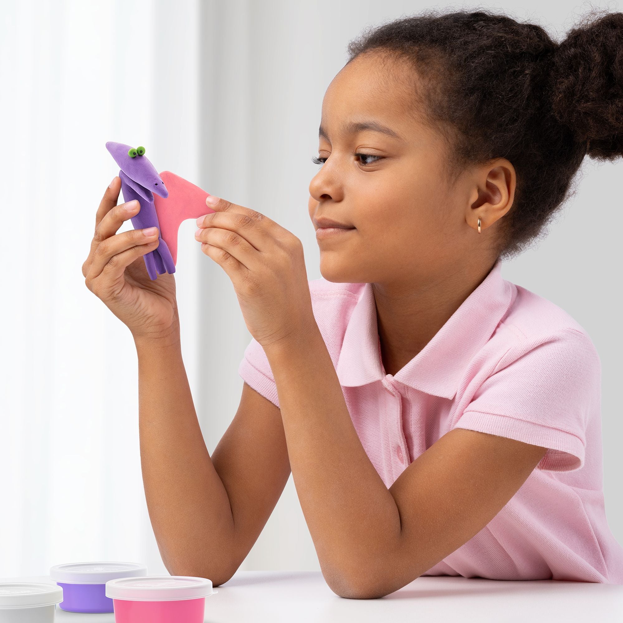 Jovem a segurar e a inspecionar uma figura de dinossauro roxo e rosa que acabou de modelar com a pasta HEY CLAY, demonstrando satisfação com o resultado.