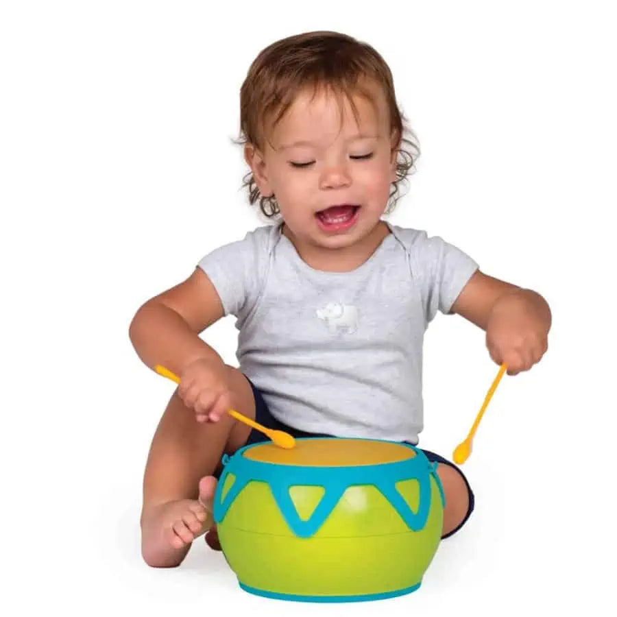 Bebé a tocar o Super Tambor Halilit sentado no chão, com expressão de concentração e alegria, segurando as baquetas.