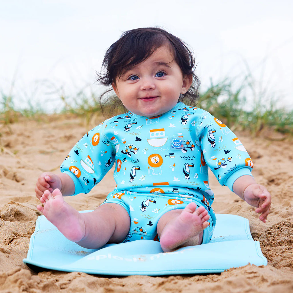 Bebé sorridente sentado na areia da praia sobre uma esteira azul, a usar o fato de praia/piscina térmico Splash About Happy Nappy Wetsuit Noah's Ark.