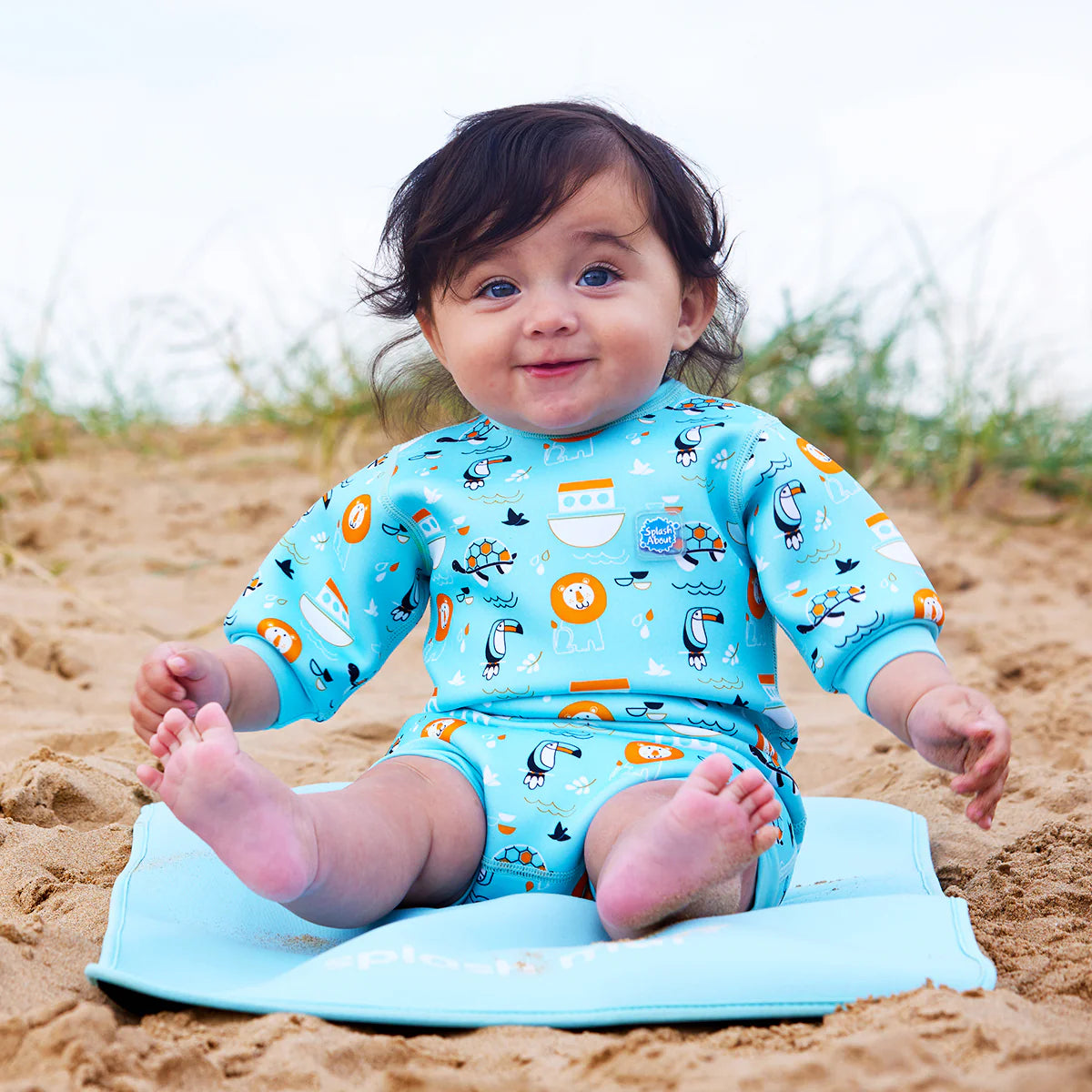 Bebé sorridente sentado na areia da praia sobre uma esteira azul, a usar o fato de praia/piscina térmico Splash About Happy Nappy Wetsuit Noah's Ark.