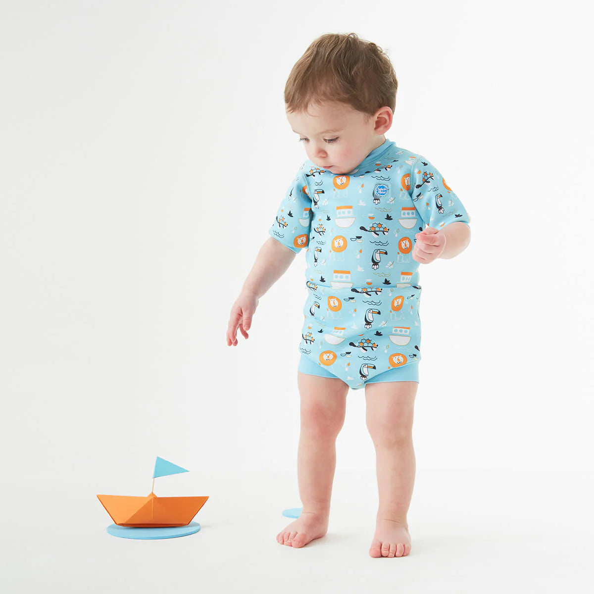 Bebé de pé, a brincar com um barco de papel, a usar o fato de praia/piscina térmico Splash About Happy Nappy Wetsuit Noah's Ark.