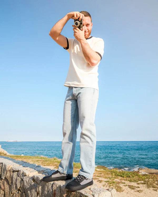 Homem em pé sobre uma rocha junto ao mar, a tirar uma fotografia com uma câmara analógica, calçando os ténis Muris Menorca.