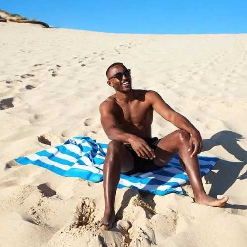 Homem sorridente sentado numa duna de areia, a usar a toalha de praia Dock and Bay azul e branca de riscas como esteira.