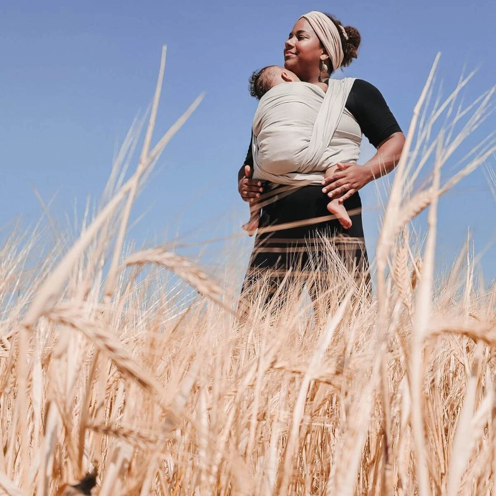 Mulher com turbante a carregar um bebé adormecido na frente com o Pano Tecido Hoppediz T6 Djerba, num campo de trigo sob céu azul.