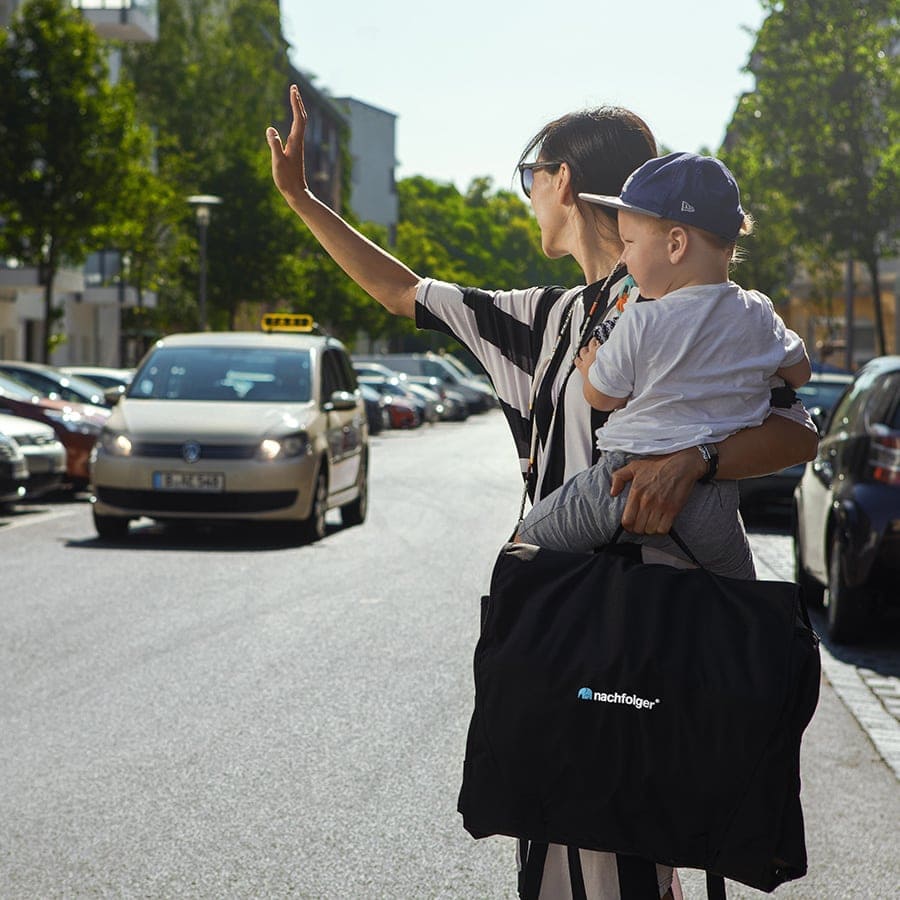 Mulher com criança ao colo a chamar um táxi, transportando a cadeira auto Nashfolger HyFive CITY R129 na mala preta, enfatizando a facilidade de uso em viagens urbanas.