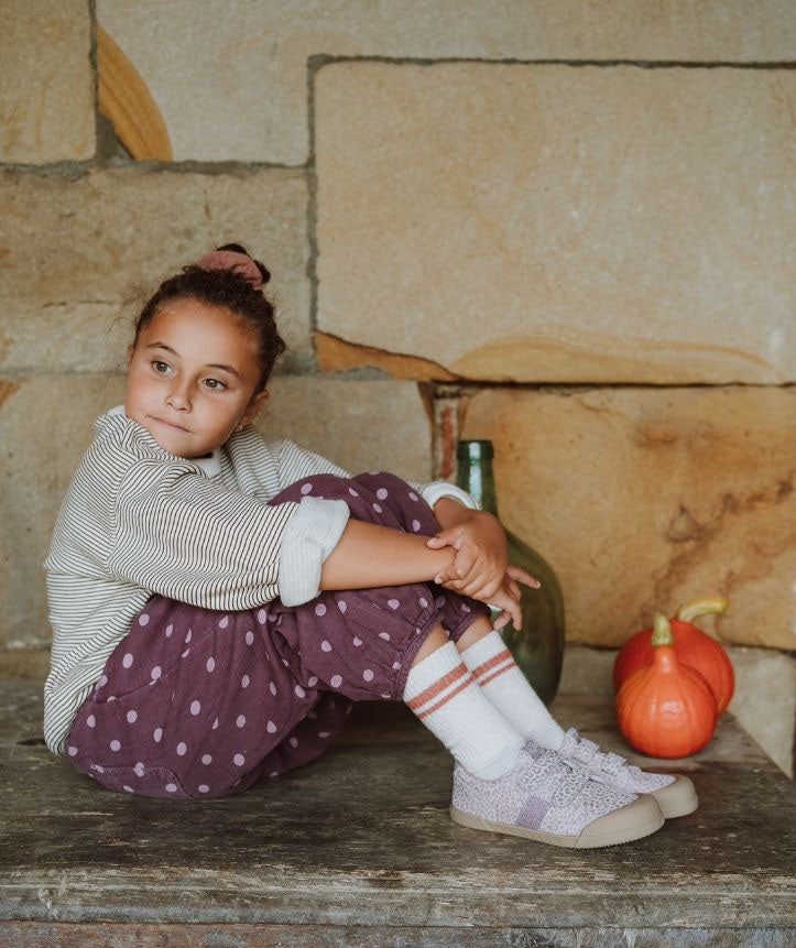 Criança sentada a usar as sapatilhas Igor Barefoot TENNIS LEO ROSA em tom lilás com padrão leopardo, em ambiente rústico.
