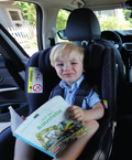 Fotografia de uma criança sentada numa cadeira auto dentro de um carro, sorrindo e segurando um livro, mostrando a praticidade e conforto do produto.