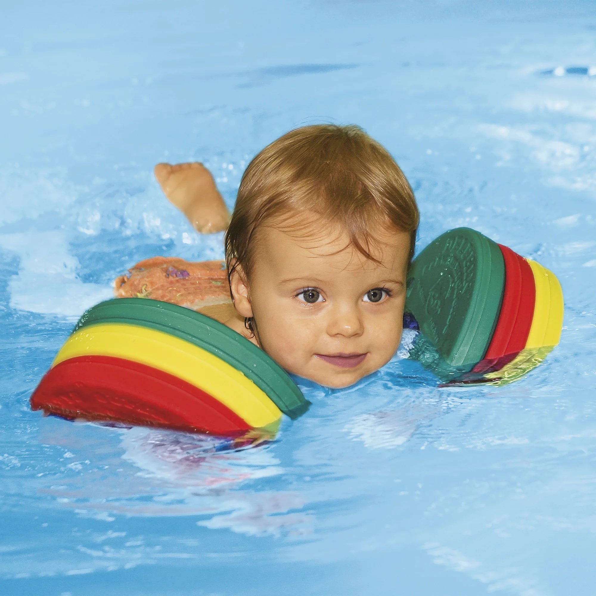 Um bebé com cabelo claro na piscina a usar as braçadeiras Delphin Discs coloridas, com uma expressão curiosa e tranquila, a flutuar na água.