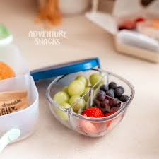Tigela de lanche Yumbox transparente com uvas, mirtilos e morangos, com uma tampa azul ao fundo.