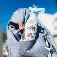 Garrafa térmica Cool Bottles Tudor Village 350ml com tampa de palhinha, inserida no bolso lateral de uma mochila cinzenta, sob luz solar.