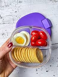Mão a segurar a tigela Yumbox com tampa roxa, contendo ovo cozido fatiado, tomate cereja e bolachas redondas.