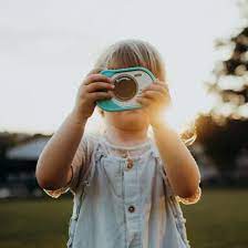 Uma criança pequena com cabelo loiro a segurar uma máquina fotográfica Kidywolf azul-esverdeada, a fotografar ao ar livre com o sol a brilhar por trás.