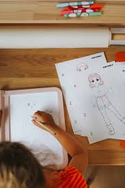 Vista aérea de uma criança a desenhar com a prancha KidyDraw rosa, com folhas de moldes numerados ao lado, focando-se na mão e no processo de desenho.