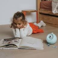 Criança deitada no chão a ler um livro aberto, com o projetor KidyStar ao lado, num ambiente calmo.