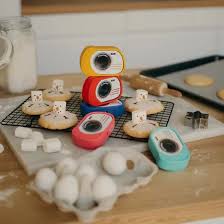 Três máquinas fotográficas Kidywolf coloridas (amarela, vermelha, azul) empilhadas, com outras câmaras e ingredientes de pastelaria espalhados numa bancada de cozinha, sugerindo brincadeira e criatividade.