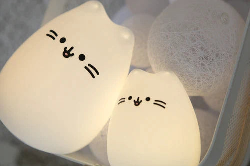 Duas luzes de presença em silicone com formato de gatinho, uma maior e outra menor, emitindo luz branca suave, rodeadas por decorações brancas.