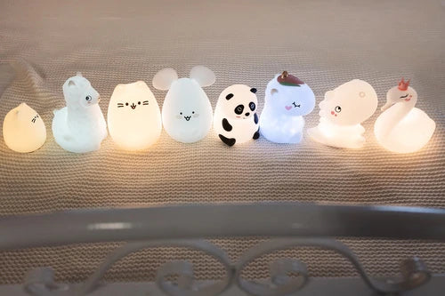 Coleção de sete luzes de presença noturnas InnoGio em silicone com formas de animais (incluindo gatinho, panda e unicórnio) alinhadas e acesas sobre uma superfície de malha clara.