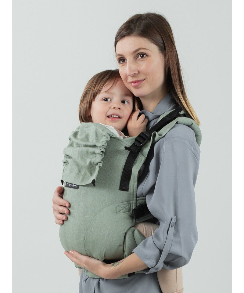 Mãe com a mochila Isara The One Sage Green Linen, carregando um bebé de cerca de 1 ano.