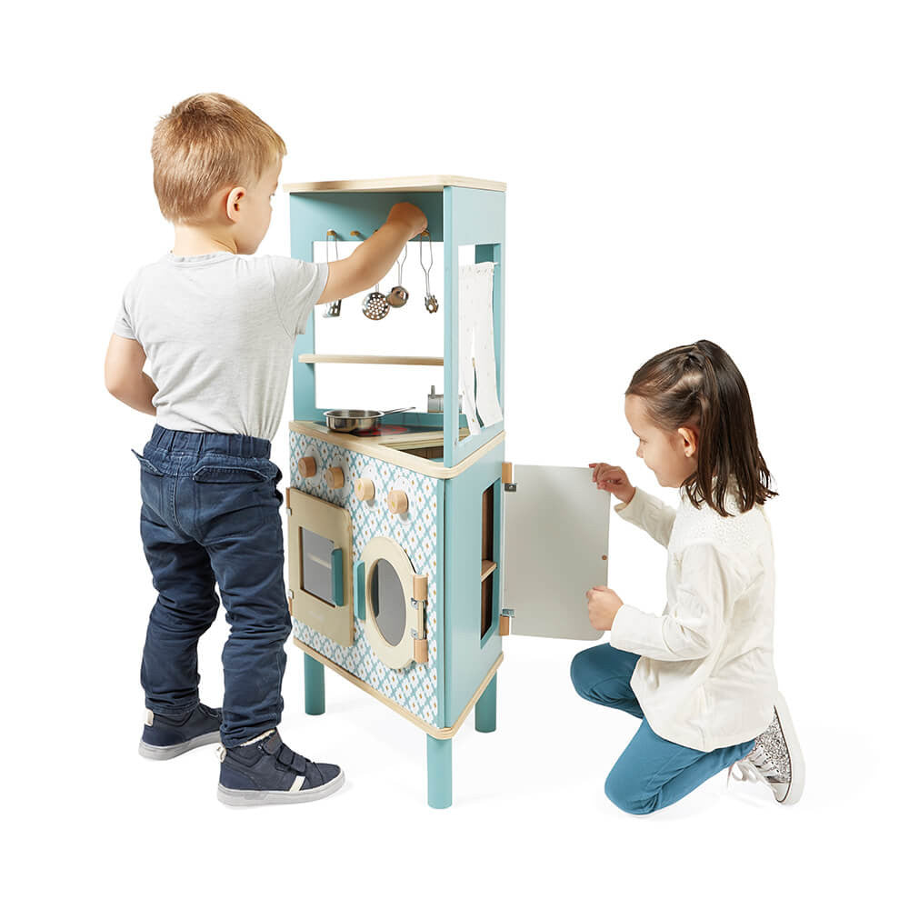 Duas crianças brincam com a cozinha triangular Janod azul; um rapaz acede aos utensílios e uma menina abre a porta da máquina de lavar.