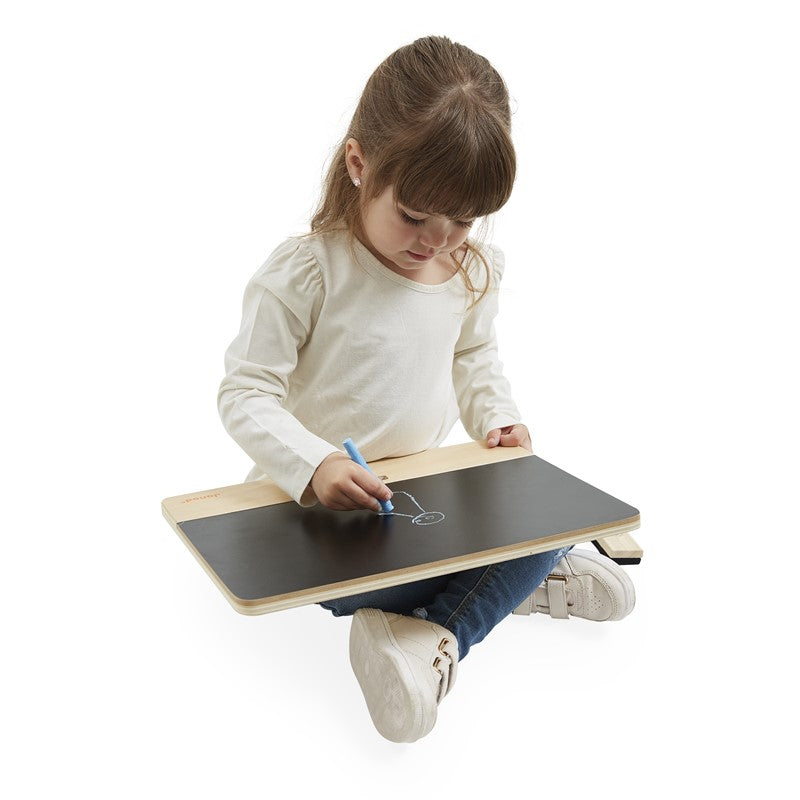 Criança sentada no chão a desenhar com giz azul numa lousa de ardósia de madeira Janod, ideal para brincar em qualquer superfície.