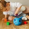 Criança pequena inclinada a brincar com o brinquedo de encaixe Narval azul, inserindo formas coloridas nos orifícios.