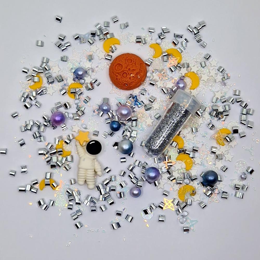 Detalhe dos elementos da recarga temática "Espaço" da Jellystone: glitter prateado, figuras de astronauta, planeta, luas e estrelas.