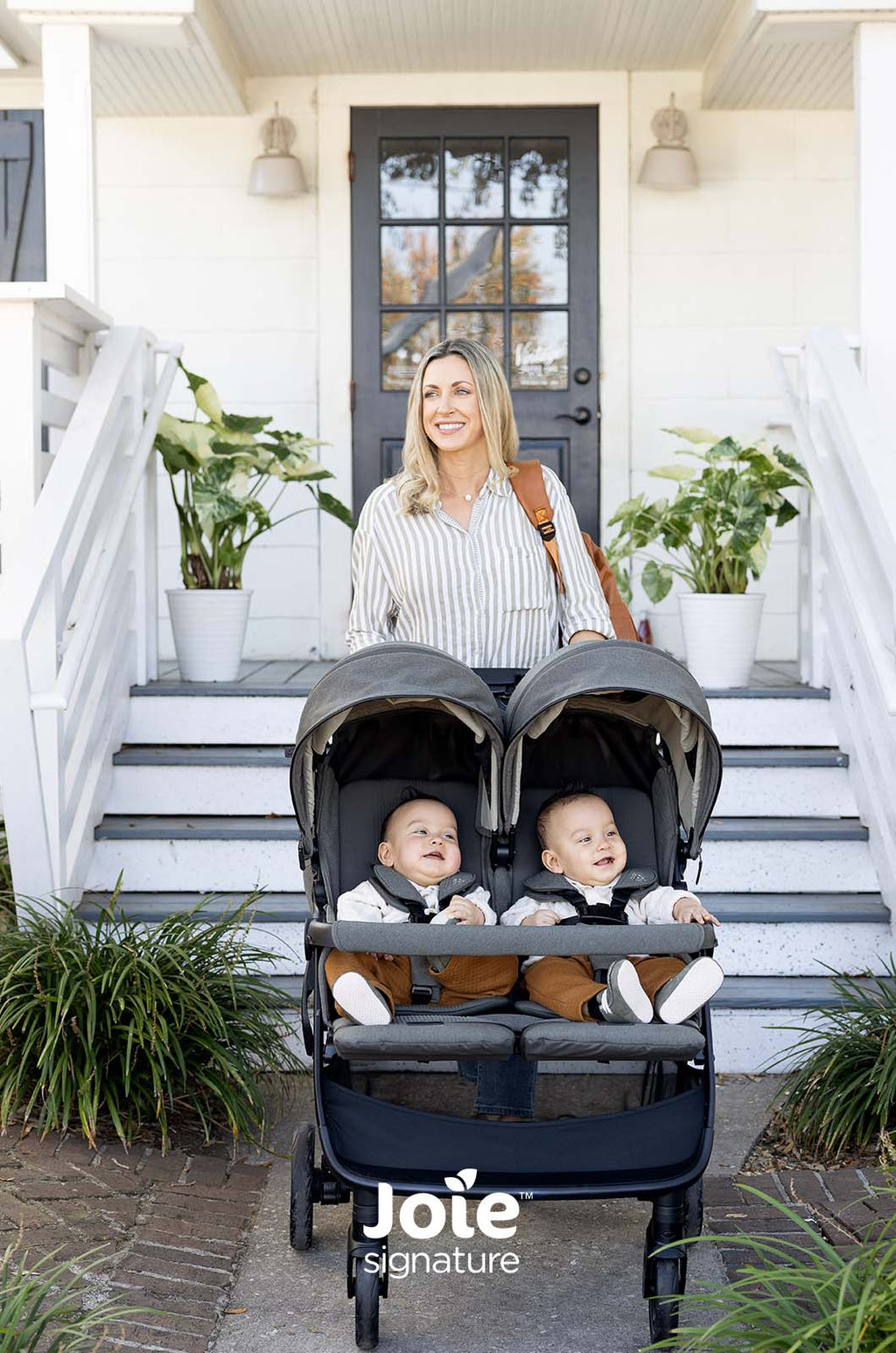 Mãe sorridente em pé atrás do carrinho duplo Joie Estrella Signature cinza, com dois bebés sentados, em frente a uma entrada de casa.