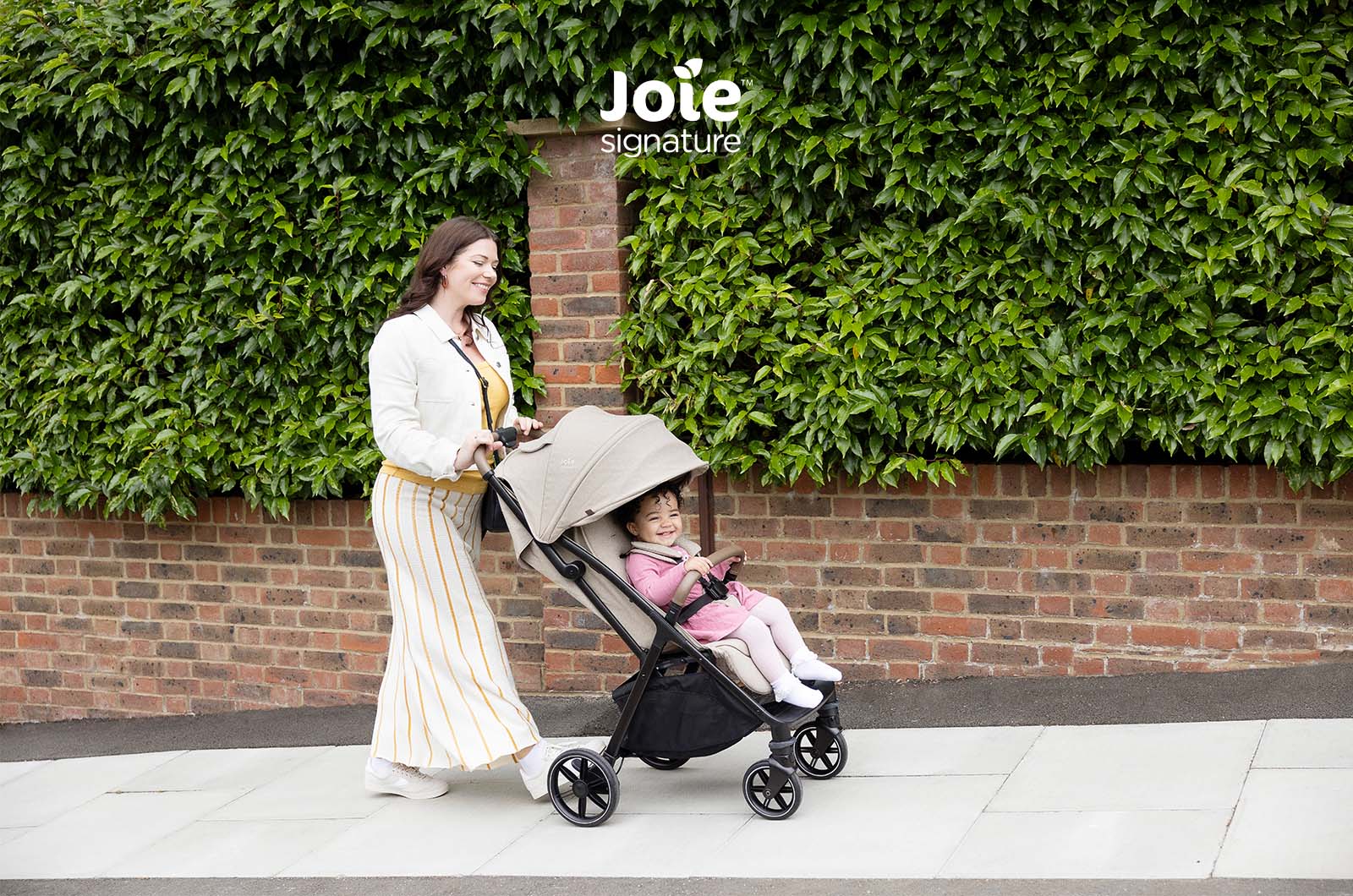 Mãe empurra o carrinho Joie Parcel LX Signature bege com criança sorridente, em passeio urbano junto a uma sebe verde.