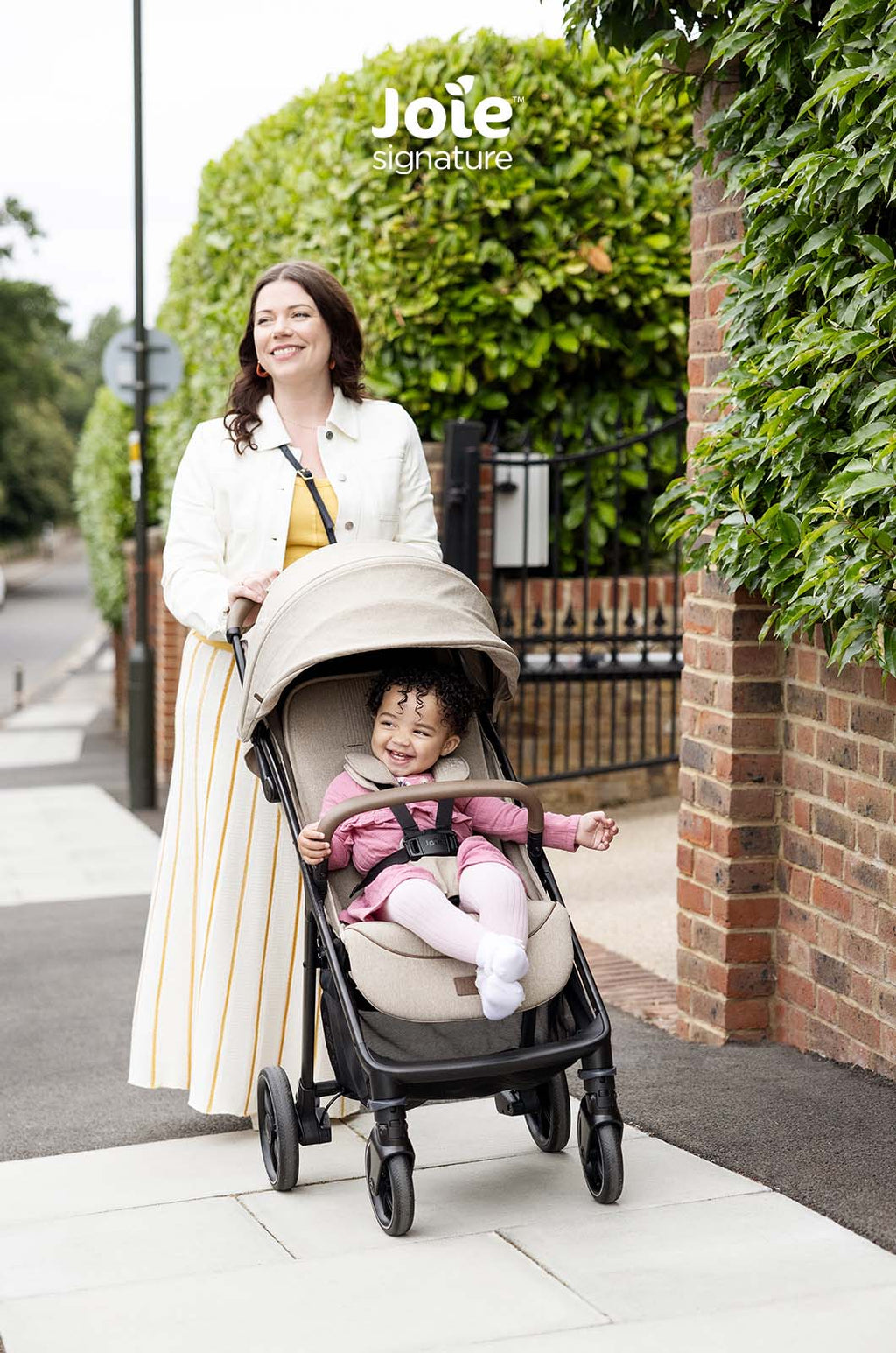 Criança feliz no carrinho Joie Parcel LX Signature, com capota estendida, enquanto a mãe o empurra num passeio junto a uma parede de tijolos.