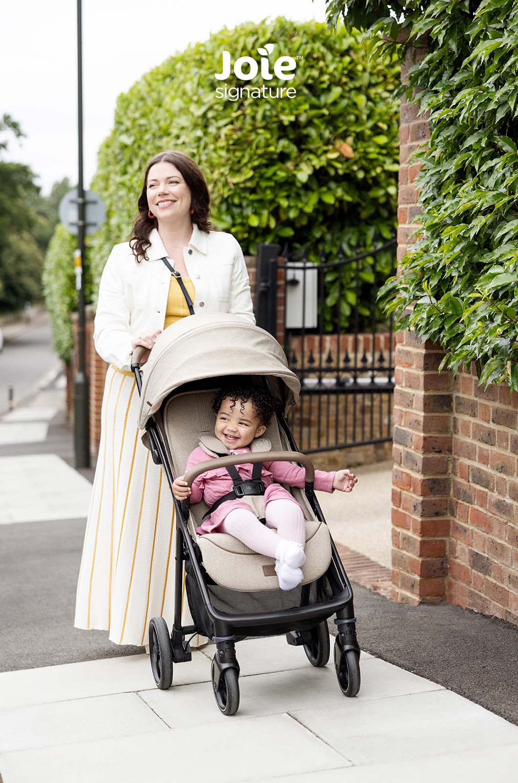 Criança feliz no carrinho Joie Parcel LX Signature, com capota estendida, enquanto a mãe o empurra num passeio junto a uma parede de tijolos.