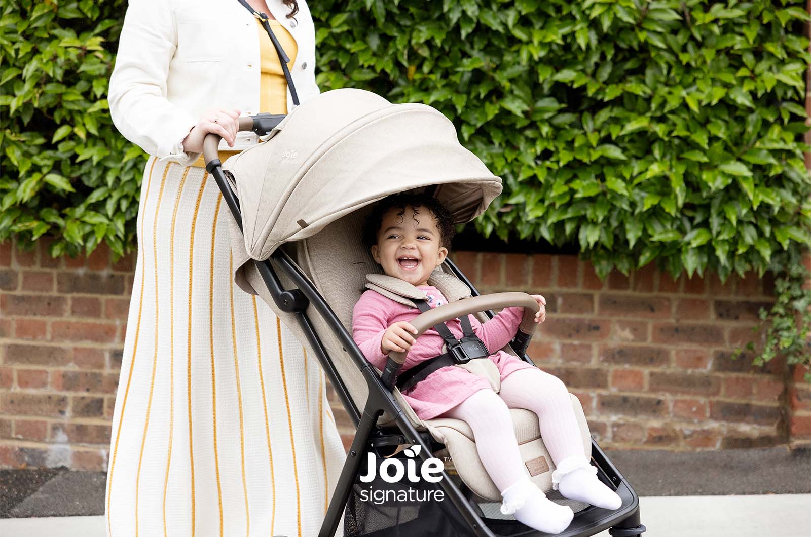 Close-up da criança sorridente no carrinho Joie Parcel LX Signature, focando no conforto do assento e arnês de segurança.