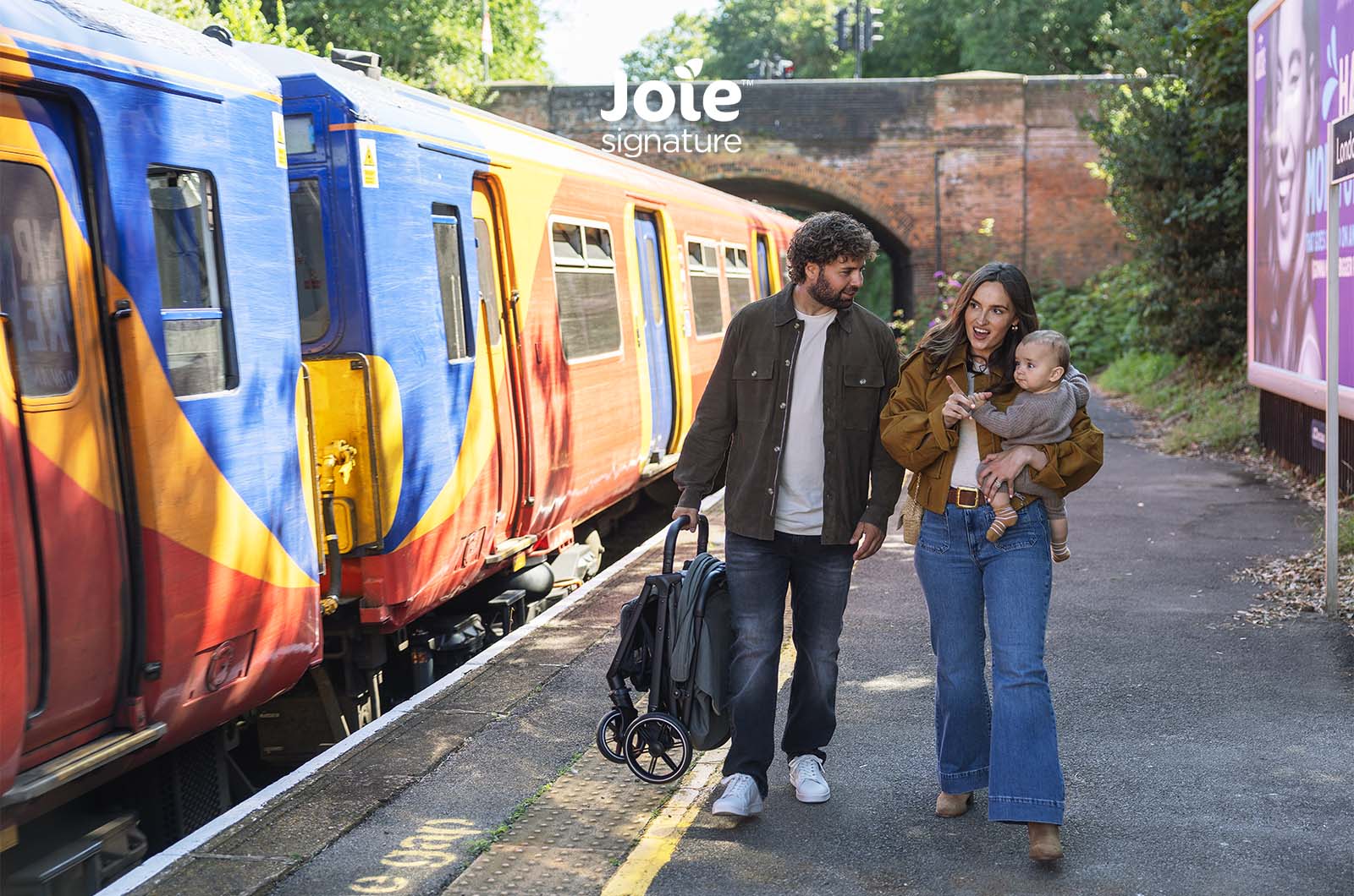 Casal com bebé numa estação de comboios, o homem transporta o carrinho Joie Parcel LX dobrado, destacando a sua portabilidade.
