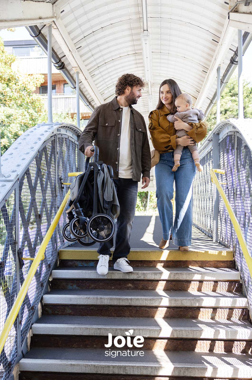 Família atravessa uma ponte pedonal, o homem carrega o carrinho Joie Parcel LX dobrado, ilustrando a facilidade de transporte em escadas.