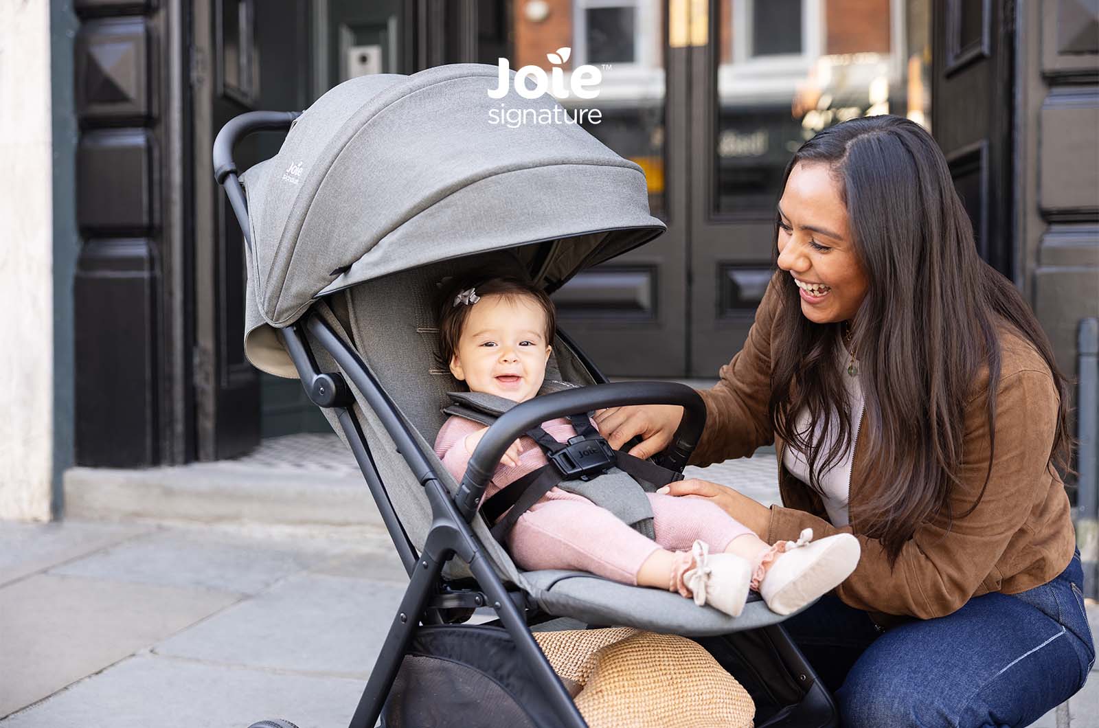 Bebé sorridente no carrinho Joie Parcel LX Signature cinzento, com a mãe a ajustar o arnês, destacando o conforto e segurança.