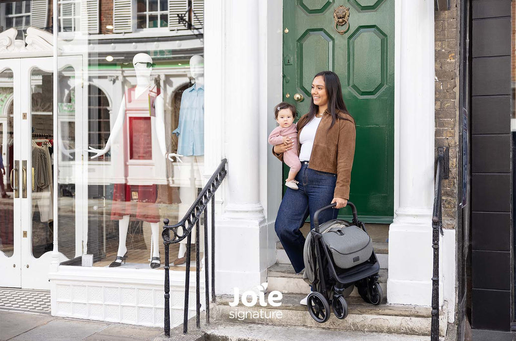 Mãe segura o carrinho Joie Parcel LX dobrado e compacto, enquanto segura o bebé, à porta de uma loja urbana.