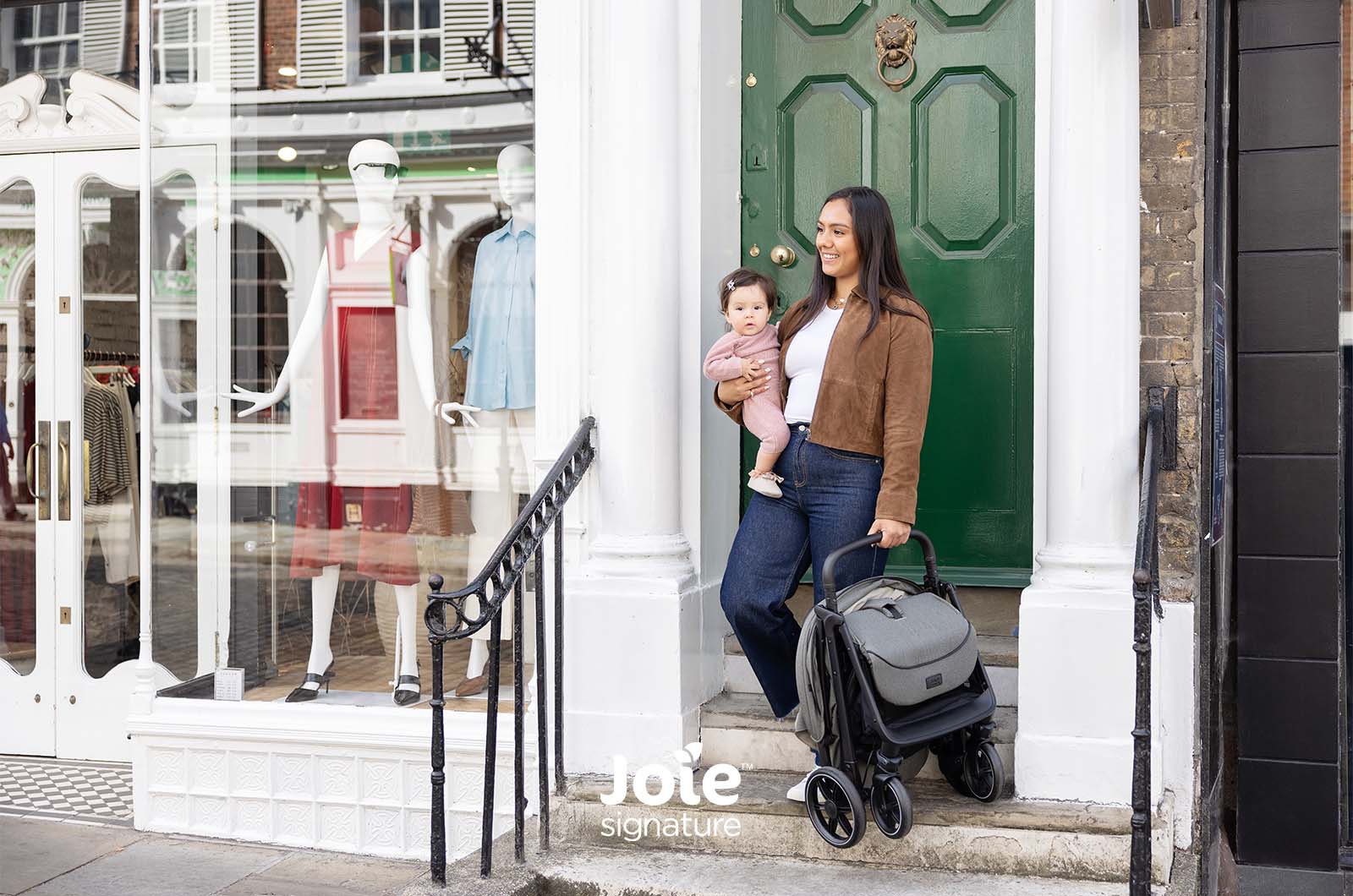 Mãe segura o carrinho Joie Parcel LX dobrado e compacto, enquanto segura o bebé, à porta de uma loja urbana.