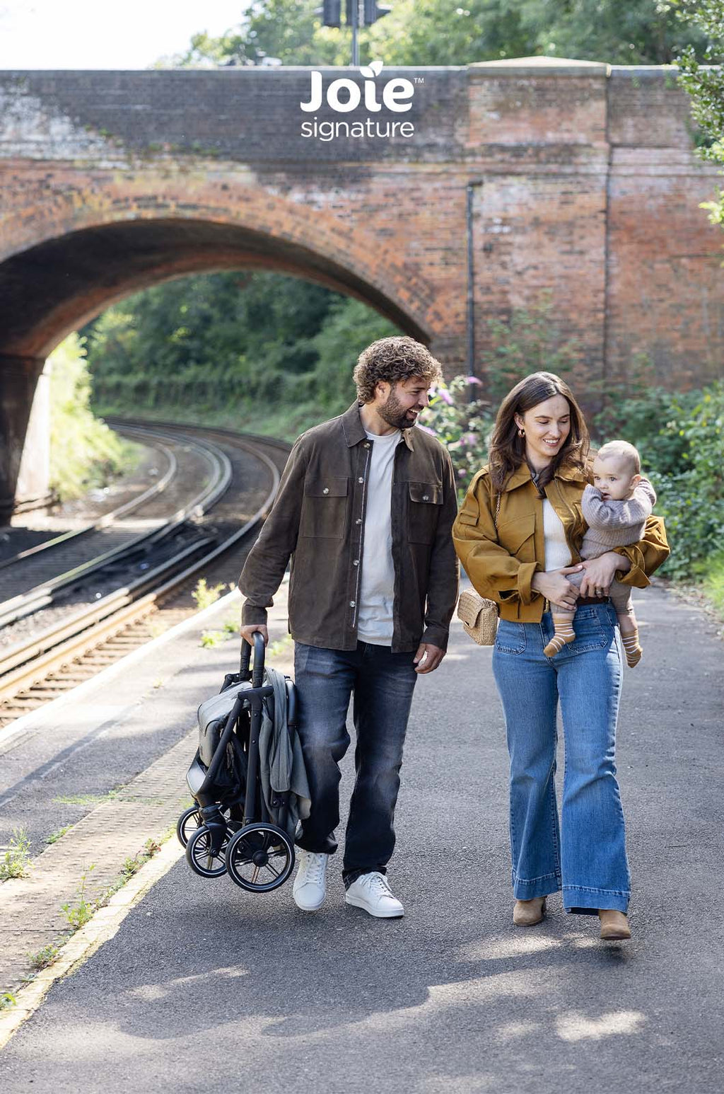 Casal caminha por uma estação de comboios, o pai transporta o carrinho Joie Parcel LX dobrado, realçando a portabilidade do carrinho.