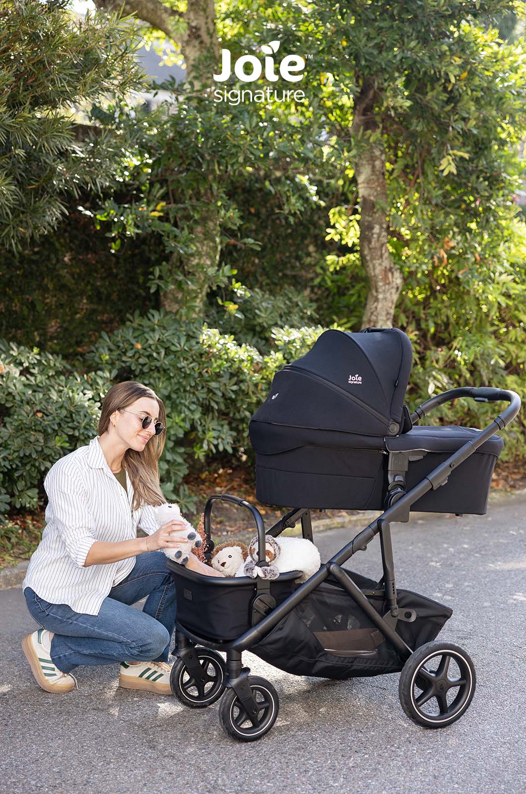 Mãe ajoelhada ao lado do carrinho Joie Versiti Signature com uma alcofa instalada, ajeitando um peluche, em ambiente exterior.