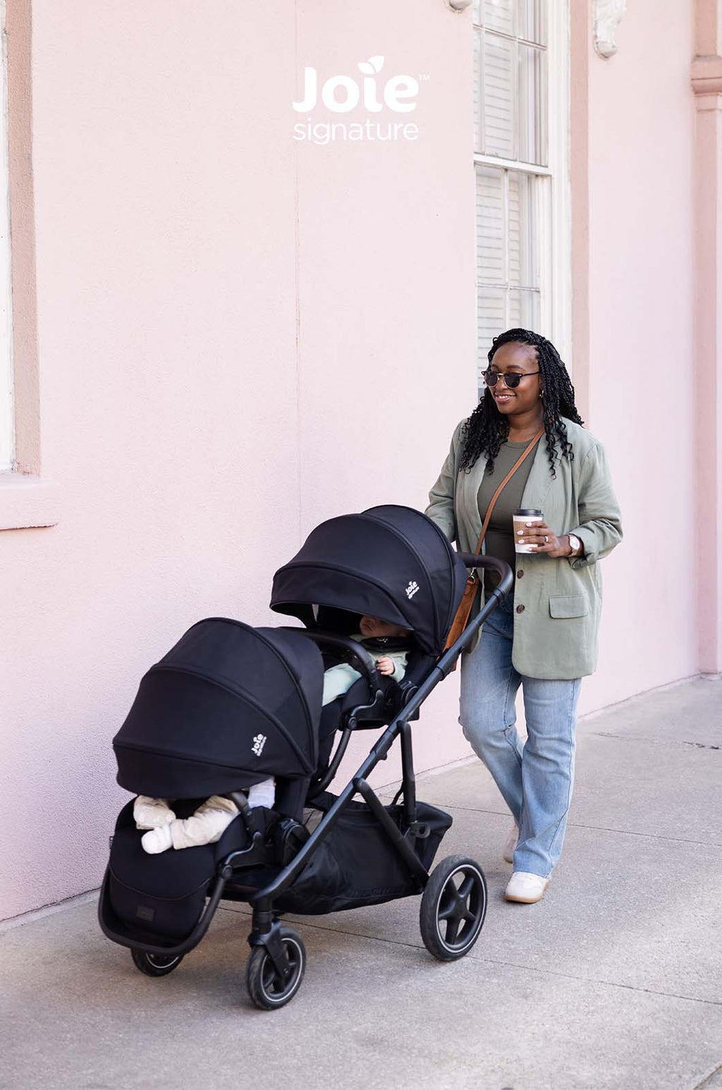 Carrinho duplo Joie Versiti Signature preto com dois bebés (um visível) a passear junto a uma parede rosa, com a mãe a sorrir.
