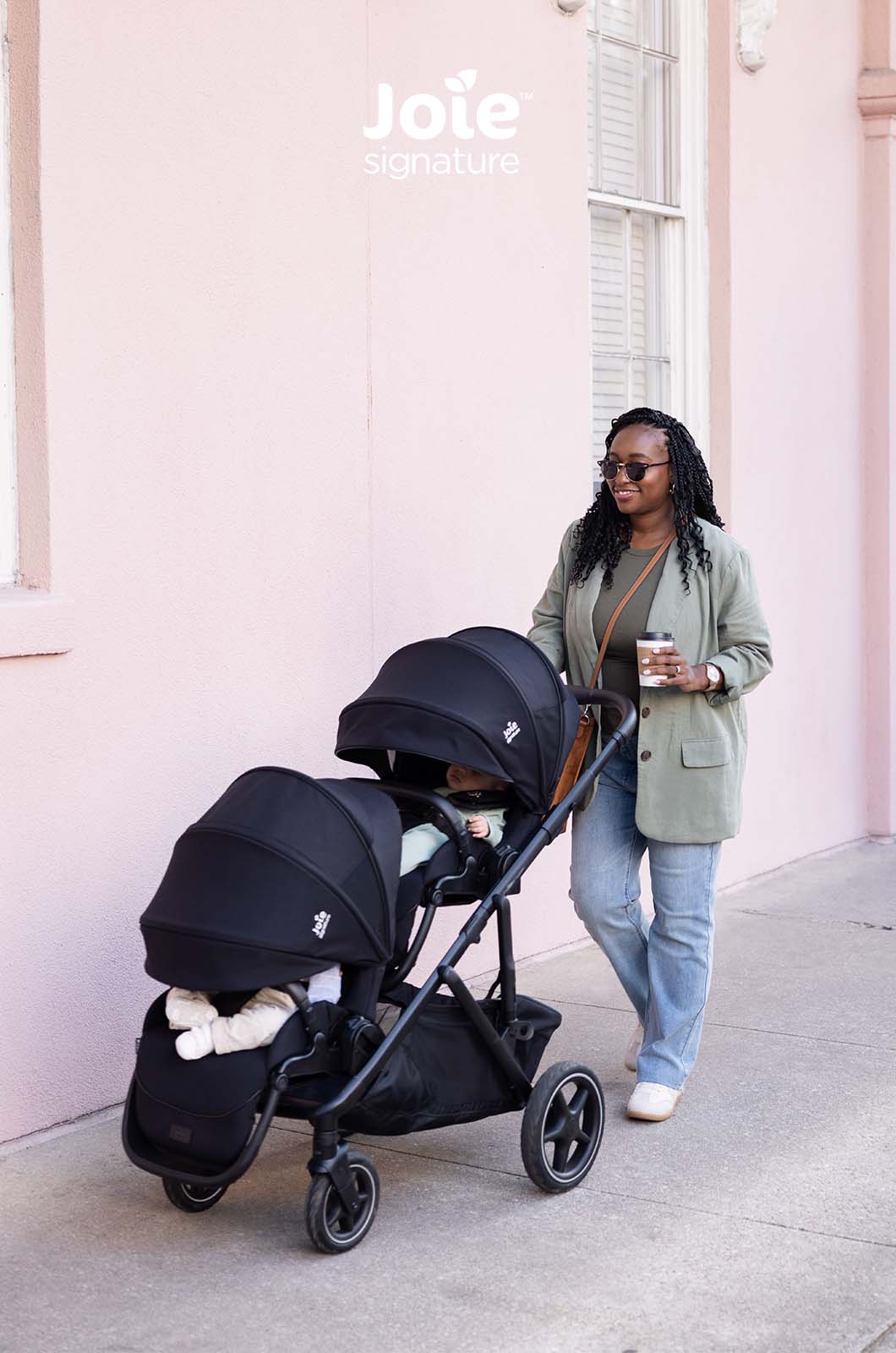Carrinho duplo Joie Versiti Signature preto com dois bebés (um visível) a passear junto a uma parede rosa, com a mãe a sorrir.