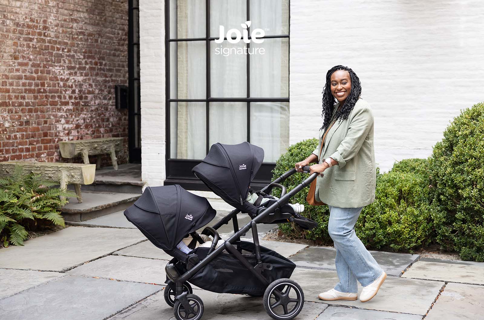 Mãe sorridente empurra o carrinho duplo tandem Joie Versiti Signature preto em passeio urbano, destacando o design elegante.