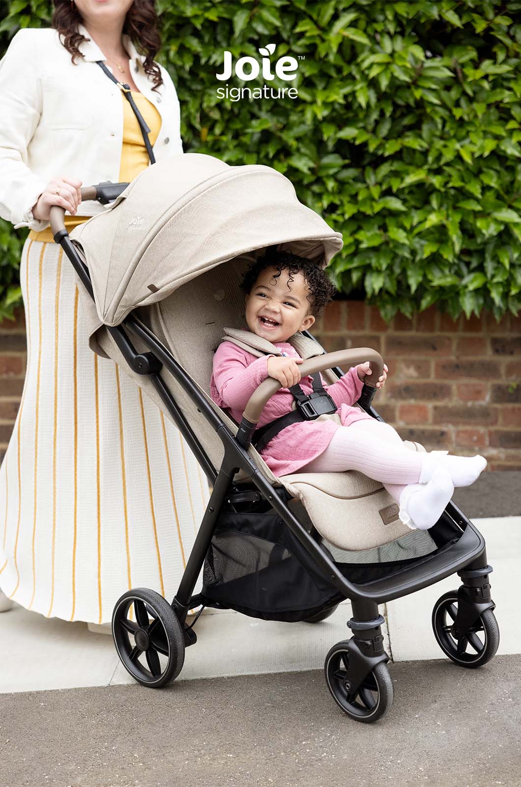 Bebé sorridente num carrinho Joie Parcel LX Signature bege, com acabamentos Signature, sendo empurrado por uma mulher em ambiente exterior.