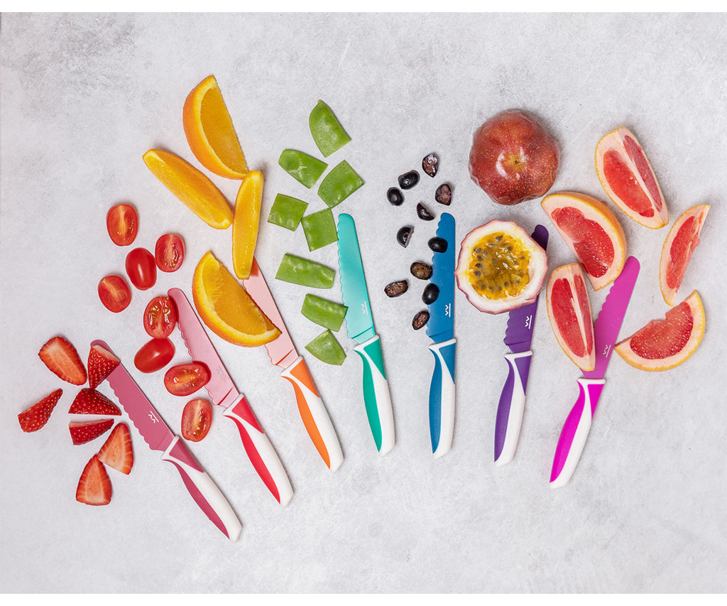 Conjunto de facas KiddiKutter coloridas (rosa, laranja, verde-água, azul, roxo) dispostas lado a lado com frutas e legumes fatiados (morangos, laranjas, pimentos, maracujá, toranja) sobre fundo cinzento claro.