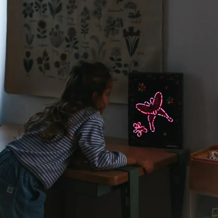 Criança a observar uma placa de borboleta iluminada com fio neon rosa num quarto escuro.
