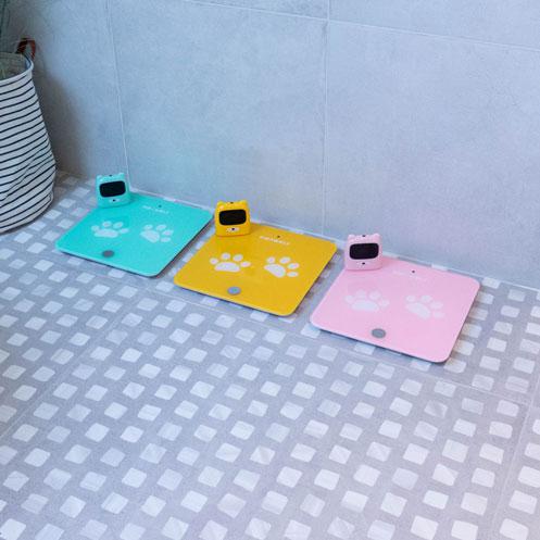 Três balanças KidyWolf (verde-água, amarela e rosa) lado a lado no chão de azulejos, mostrando as opções de cor disponíveis.